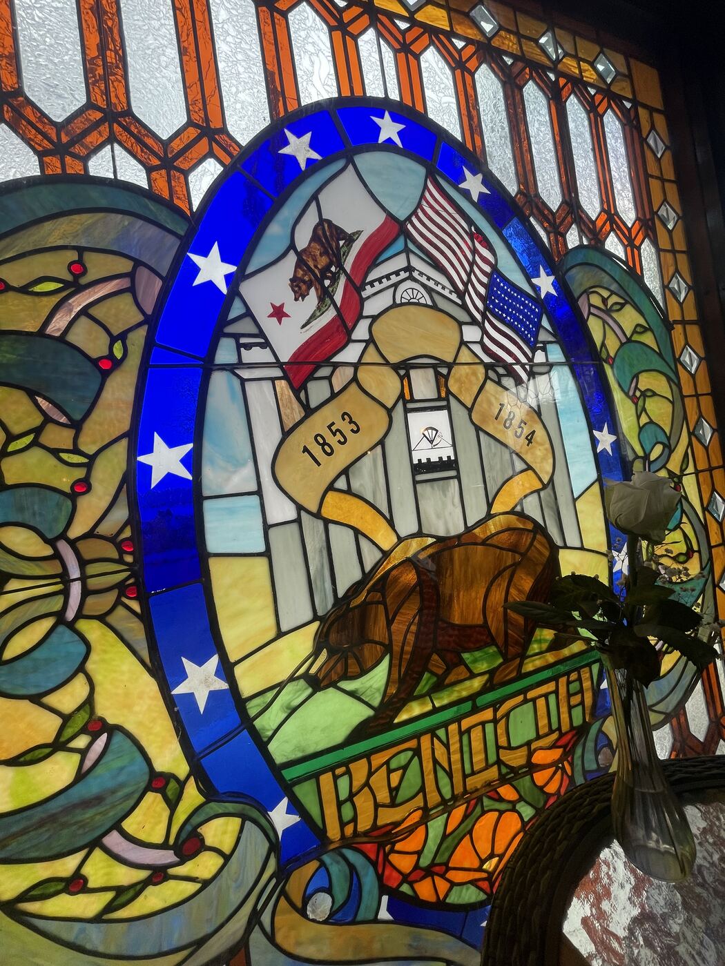 A stained glass window reading Benicia, showing the old Capitol building, the California bear flag and the United States flag, and ribbons marking 1853 and 1854.