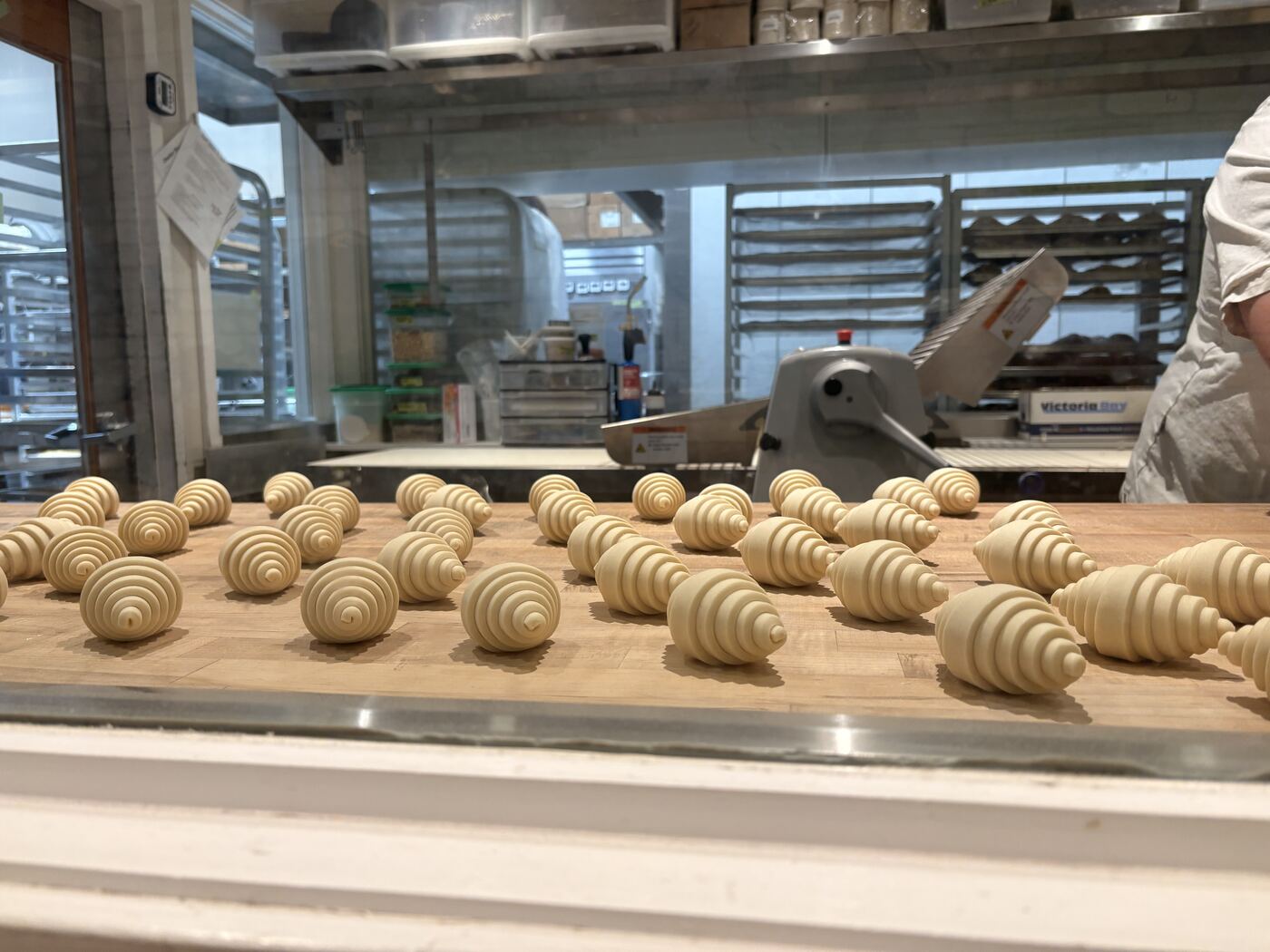 Rows of hand-rolled croissant dough resting on a wooden bench in a bakery, with the kitchen and a baker at work behind the glass.