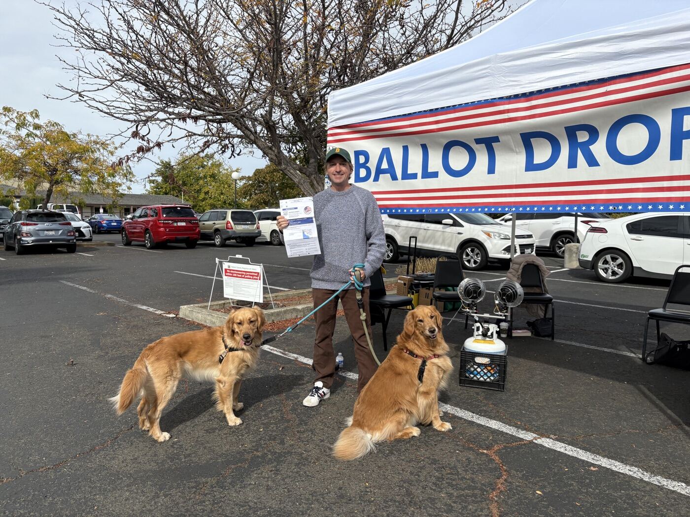 Justin Zollars holding his ballot beside a county Ballot Drop-off tent, with two golden retrievers on leash.